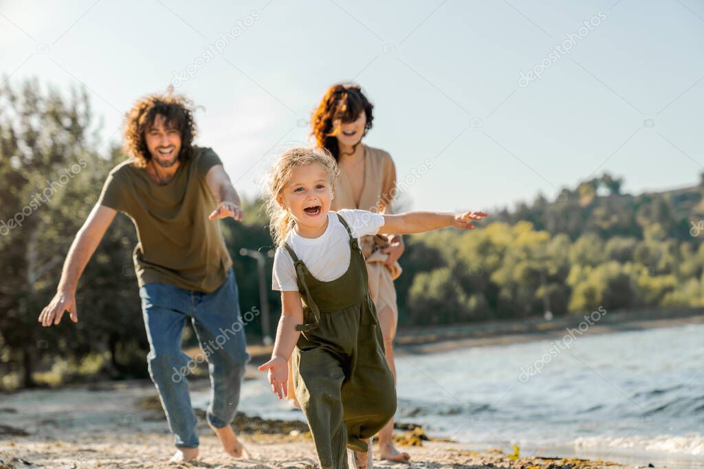 Happy young family having fun on the beach. Little cute girl running along a beach being chased by her parents.