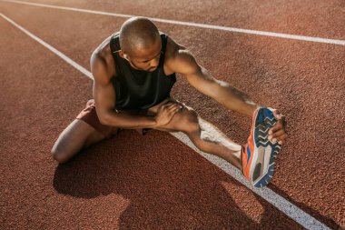 Erkek koşucu, stadyumda otururken ayakkabısına dokunarak bacak kaslarını esnetiyor.