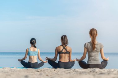 Arka planda, nilüfer çiçeğiyle yoga yapan kadınların deniz kenarında güneşli plajda poz verirken görülüyor.