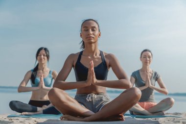 Spor salonundaki üç kadın yoga ve meditasyon yaparken deniz kıyısında oturuyorlar..