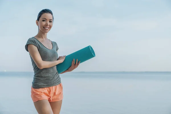 Smiling asian female athlete in fitness clothes relaxing after workout with mat in hand standing on the beach