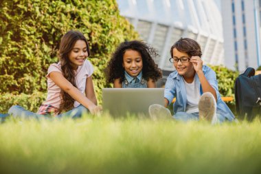 Dijital aletleri ve sırt çantalarıyla dışarıdaki çim parkında oturan mutlu küçük okul çocuklarının düşük açılı görüntüsü.