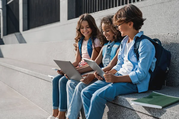 Счастливая Группа Мультиэтнических Школьников Сидящих Пользующихся Цифровыми Устройствами Улице — стоковое фото
