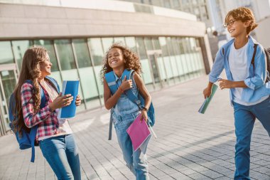 Okul binasının yakınında sırt çantalı mutlu çocuklar birlikte yürüyüp gülüyorlar.