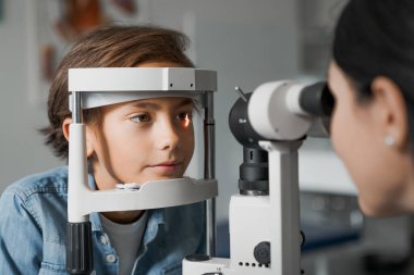 Yetişkin kadın doktor göz doktoru, modern klinikteki sevimli çocuğun bakışlarını kontrol ediyor.