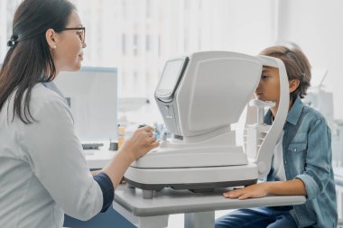 Küçük çocuğun, kadın doktorun özel optik aparatıyla gözleri için test çekerken yan görüntüsü.