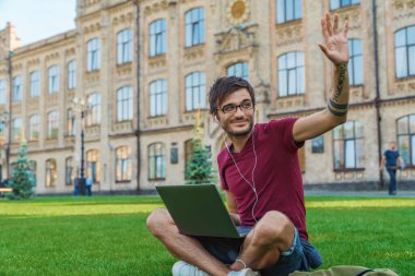 Genç bir işadamı ya da öğrenci, gözlüklü, kulaklıklı, şehir parkında oturuyor, bilgisayarını kullanıyor ve merhaba işareti yapıyor.