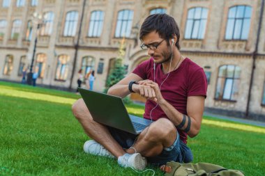 Dışarıda otururken saatine bakarken dizüstü bilgisayarda çalışan çok ırklı yakışıklı bir adam.