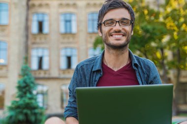 Gülümseyen öğrenci kahve molası veriyor. Genç yakışıklı adam şehirde dizüstü bilgisayar kullanıyor..