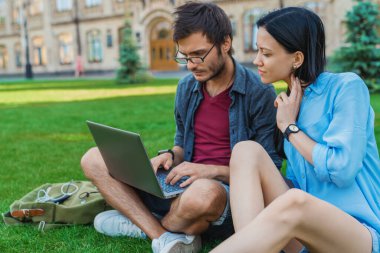 Üniversitenin yakınındaki çimlerde otururken dizüstü bilgisayar kullanan sıradan giysili hoş bir çift.