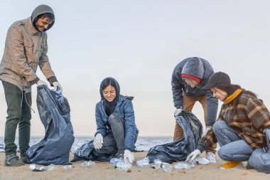 Genç adam plajda bir grup gönüllüyle çöp ve plastik topluyor.