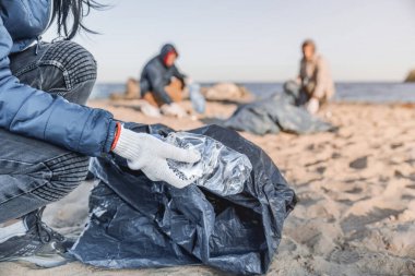Plajda arkadaşlarıyla plastik şişe toplayan eldivenli genç bir kızı yakından çek.