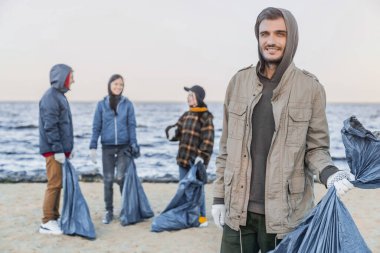 Plajda arkadaşlarıyla plastik çöp topladıktan sonra kameraya gülümseyen genç bir erkek gönüllü portresi.