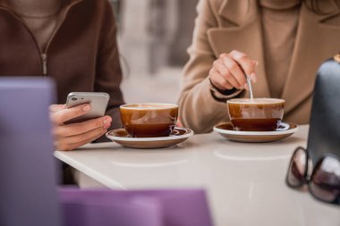 Açık kafede alışveriş yaptıktan sonra akıllı telefon kullanan ve kahve içen genç kadınların görüntüsü.