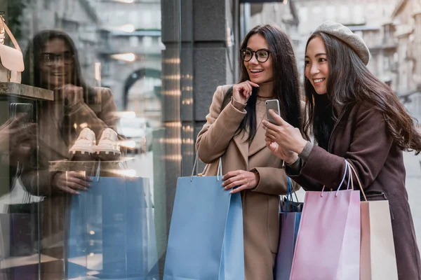Két Boldog Barátnő Nézi Kirakatot Miközben Fényképeznek Okostelefonon Bevásárlószatyrokkal Bevásárlóközpont — Stock Fotó