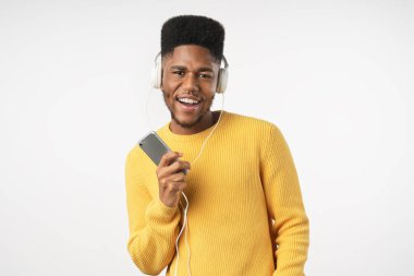 Genç Afrikalı bir adam beyaz arka planda izole edilmiş kulaklık ve cep telefonuyla müzik dinlerken şarkı söylüyor.