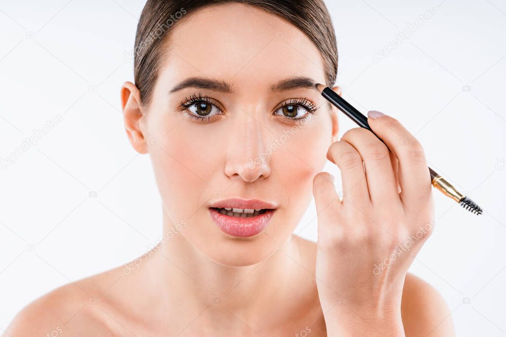 Close up studio shot of beautiful young woman applying makeup to her eyebrow while standing on isolated white background