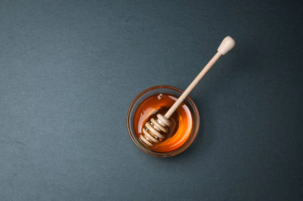 Μικρό γυάλινο μπολ με φυσικό μέλι με το ραβδί. Στον πίνακα μαύρη πέτρα ρουστίκ. — Φωτογραφία Αρχείου