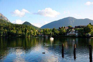 Gölün kıyısındaki evler ve rıhtımlar. Arkasında orman olan dağlar. Güzel, berrak gökyüzü, akşam aydınlığı. Gölde beyaz bir tekne.