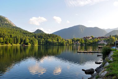 Gölün kıyısındaki evler ve rıhtımlar. Arkasında orman olan dağlar. Güzel açık gökyüzü, akşam aydınlığı.