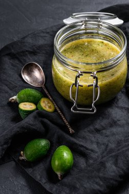 Cam bir kavanozda, tahta kesme tahtasının üzerinde şekerli, öğütülmüş yeşil feijoa. Siyah arka plan. Üst görünüm