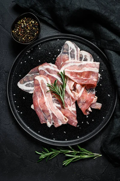 Rå bacon på svart tallrik, peppar och rosmarin. Ekologiskt kött från jordbruket. Svart bakgrund. Ovanifrån — Stockfoto