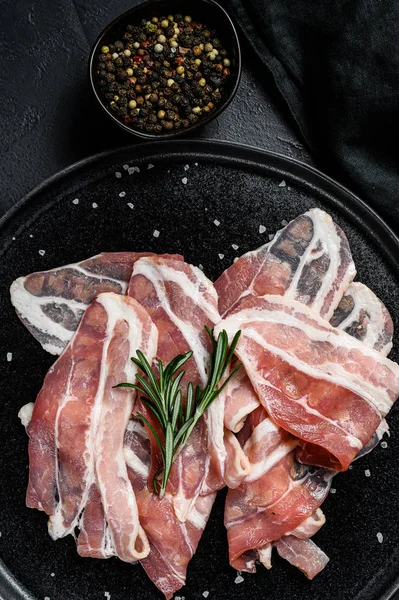 Rå bacon på svart tallrik, peppar och rosmarin. Ekologiskt kött från jordbruket. Svart bakgrund. Ovanifrån — Stockfoto