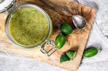 Cam bir kavanozda, tahta kesme tahtasının üzerinde şekerli, öğütülmüş yeşil feijoa. Gri arka plan. Üst görünüm.