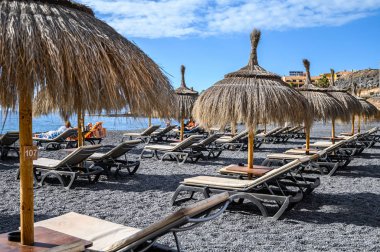 Mavi gökyüzü olan saman plajı şemsiyesi La Caletta 'nın siyah volkanik plajında güneşli yataklar. Tenerife, Kanarya Adaları, İspanya.