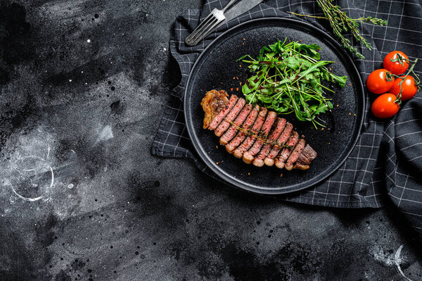 Cooked grilled strip loin steak, marbled beef meat with arugula. Black background. Top view. Copy space