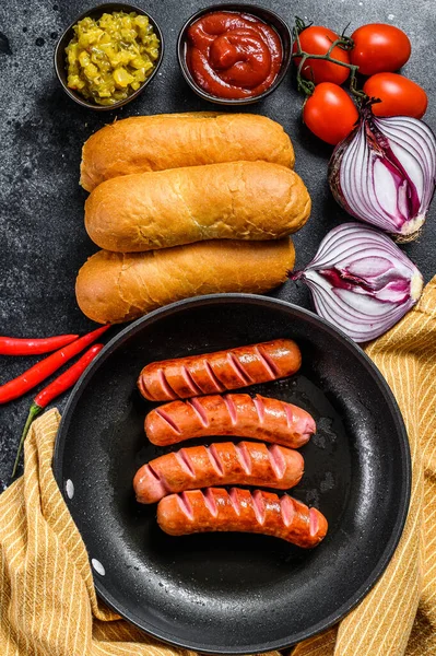 Ingredients for making homemade hot dogs. Sausages in pan, fresh baked buns, mustard, ketchup, cucumbers. Black background. Top view — Stock Photo, Image