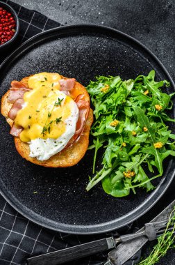 Pastırmalı Brioche sandviç, Benedict usulü yumurta ve Hollandaise sosu. Arugula salatalı garnitür. Siyah arka plan. Üst görünüm.