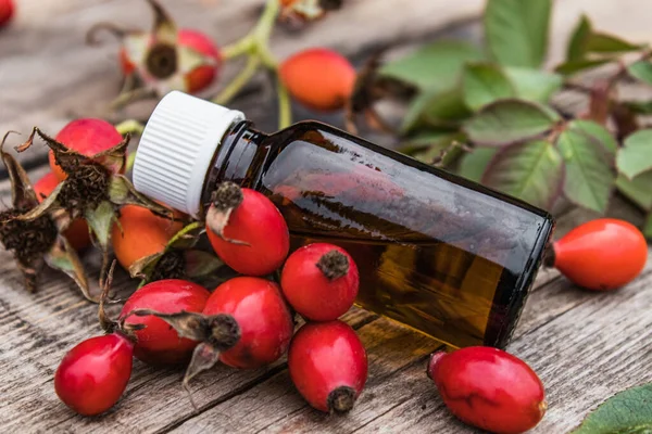 熟した赤いバラの果実の近くの木製のテーブルの上にエッセンシャルオイルを浸透させます。バラの腰でチンキやエッセンシャルオイル. — ストック写真