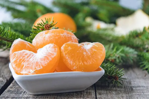 Slices of ripe yellow mandarin lie in a bowl next to the green branches of a Christmas tree. — Stock Photo, Image