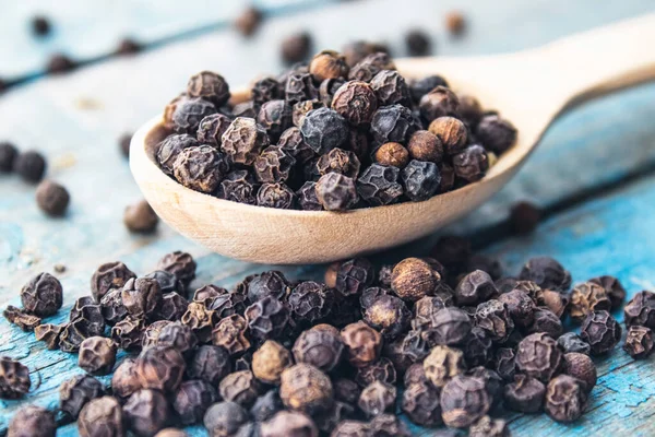 Dry black pepper in a wooden spoon near black pepper scattered on blue boards. Black pepper spices and seasonings for cooking.