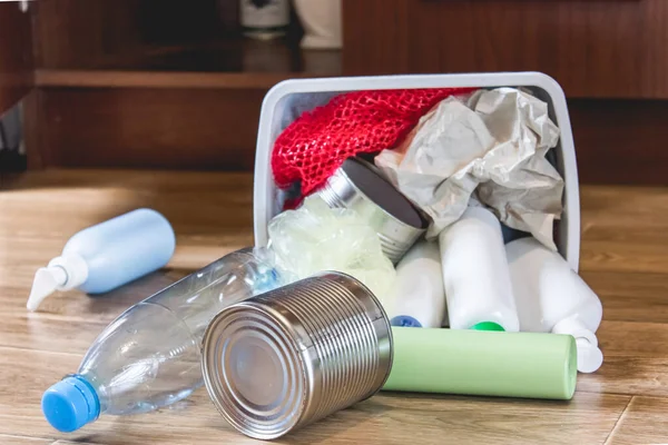 A plastic trash can with garbage lies on the floor near the kitchen cabinet - bottles, plastic and metal cans scattered in the kitchen. Garbage waste.