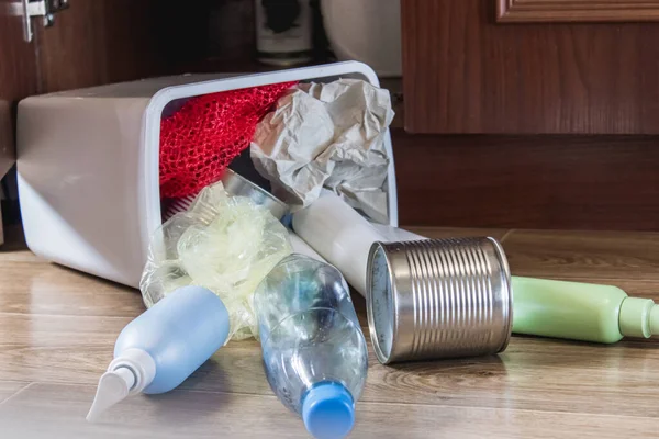 A plastic trash can with garbage lies on the floor near the kitchen cabinet - bottles, plastic and metal cans scattered in the kitchen. Garbage waste.