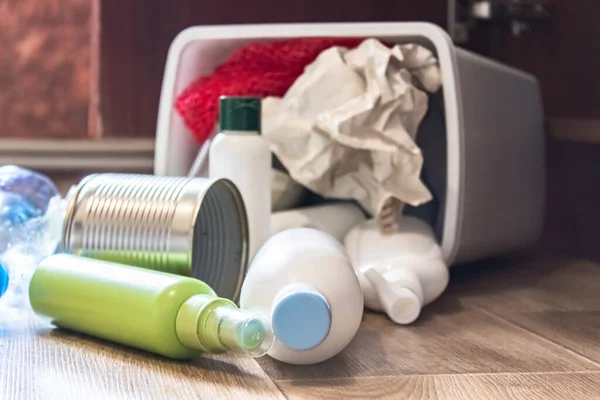 A plastic trash can with garbage lies on the floor near the kitchen cabinet - bottles, plastic and metal cans scattered in the kitchen. Garbage waste.