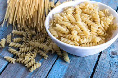 Farklı bir makarna yığınının yanındaki mavi tahtaların arka planında bir kasede kuru makarna. Sağlıklı beslenme ya da sağlıklı yaşam tarzı.