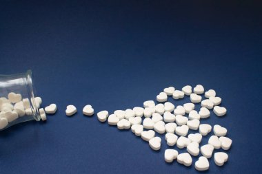 Many of white candy heart-shaped pills spilled out of a transparent jar on a dark blue background. in heart shape