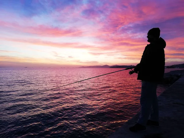 Silhouet van een mannelijke visser op de zeedijk met stenen, die aan het vissen is op de prachtige kleurrijke zonsondergang. — Stockfoto
