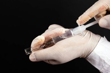 Doktor, siyah arka planda beyaz steril eldivenlerle ampulden şırıngaya ilaç topluyor.