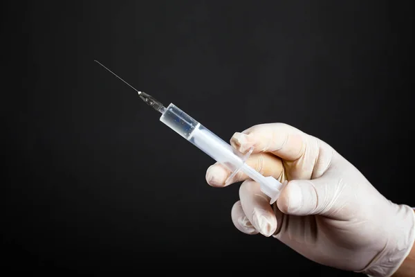 Hand in een wegwerp steriele witte handschoen met een medische spuit van 5 ml op een donkere achtergrond close-up. een injectie behandeling schoot — Stockfoto