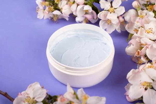 body scrub in a white jar with white apple flowers on a purple background  close-up. beauty, spa, skin care, cosmetics