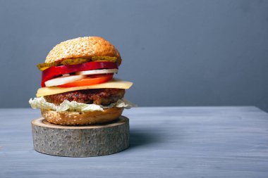 Büyük, sulu bir burger, yumuşak çörek peyniri taze sebzeler ve lezzetli et pirzolası, arka plandaki gri fotokopi alanının üzerinde, ahşap bir standın üzerinde.