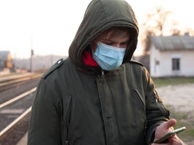 Adam istasyonda tek kullanımlık tıbbi bir maskeyle duruyor. Karantina nedeniyle toplu taşıma ve sınırın telefonunu elinde tutuyor.