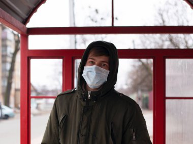 Tıbbi maskeli bir adam otobüs durağında duruyor. Viral hastalıklardan korunmak için izole edilmiş. Coperid-19. Kızamık gribi. Suçiçeği.