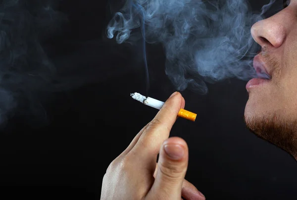 Mladý muž vydechuje kouř. poškození z kouření cigarety tmavé pozadí close-up — Stock fotografie