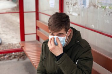 Genç adam bir otobüs durağında tek kullanımlık tıbbi bir maskeyle oturuyor. Viral solunum hastalığına bağlı karantina covid-19. 