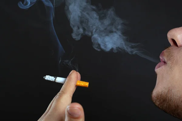Jovem segurando um cigarro fumando em suas mãos e exala fumaça em um fundo escuro. danos causados pela nicotina e tabaco close-up — Fotografia de Stock